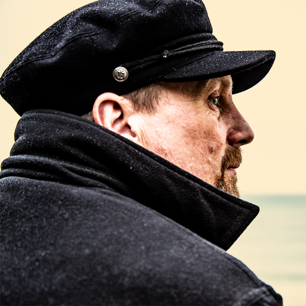 Sam Rogers in a fisherman's cap and a high collared coat looking to the right