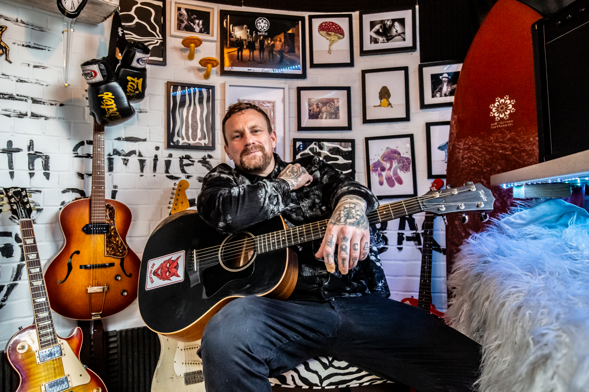 Sam rogers sat in a comfy chair with a black acoustic guitar across his lap with his arms resting over it in front of a wall of multiple pieces of framed art, more guitars and boxing gloves.